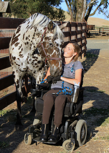 A smiling person in a wheelchair sharing a tender moment with a dappled horse. They are outdoors, possibly at a ranch, with wooden fences in the background suggesting an equestrian setting. The sunlight highlights the affectionate exchange, underscoring a bond between human and animal.