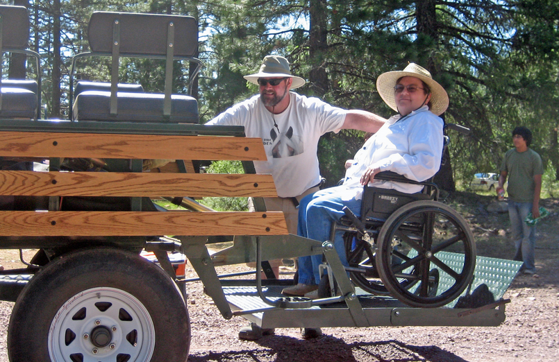 In this image, a wheelchair-accessible vehicle is shown in an outdoor setting. A person in a wheelchair is situated on a specially designed platform of the vehicle, with another individual standing beside, perhaps assisting or ready to accompany them. Both wear hats, suggesting readiness for an outdoor excursion, while another person is visible in the background, contributing to the communal, inclusive atmosphere of the setting.